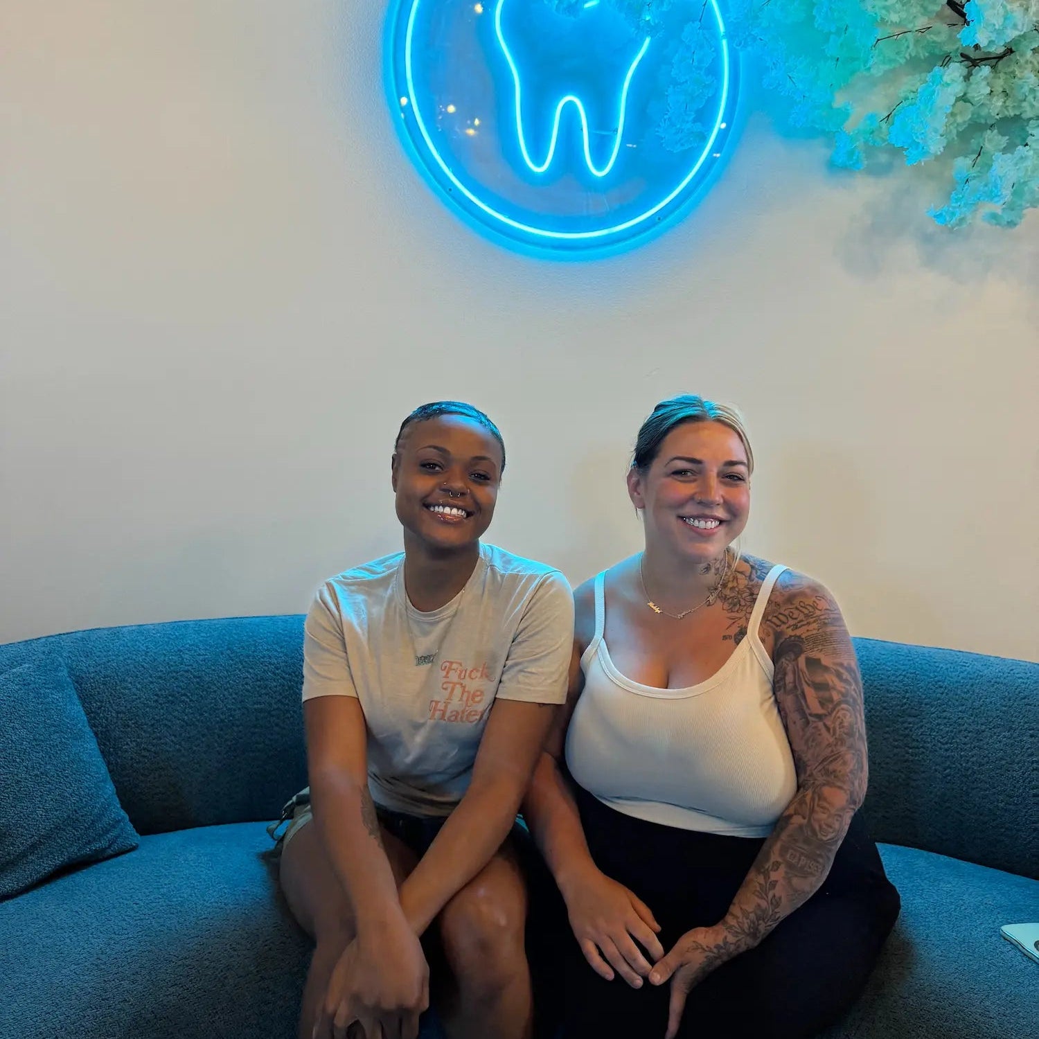 Two people sitting on a blue couch with a neon tooth sign in the background. grin & glow 