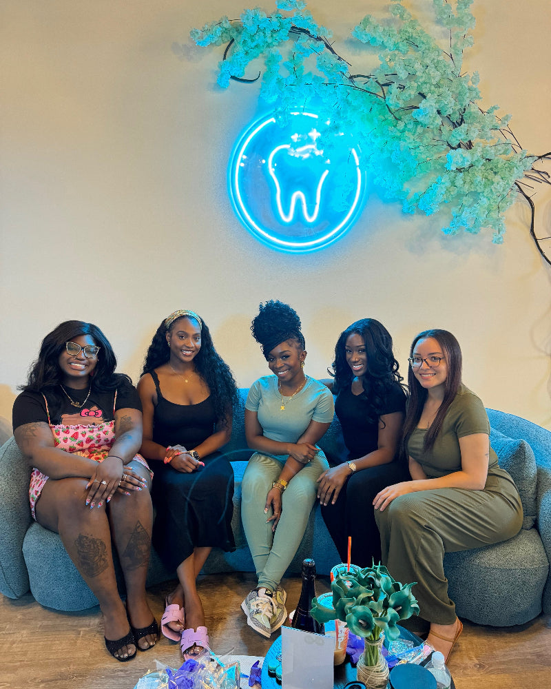 Five women sitting on a couch in a room with a neon tooth sign and floral decorations.