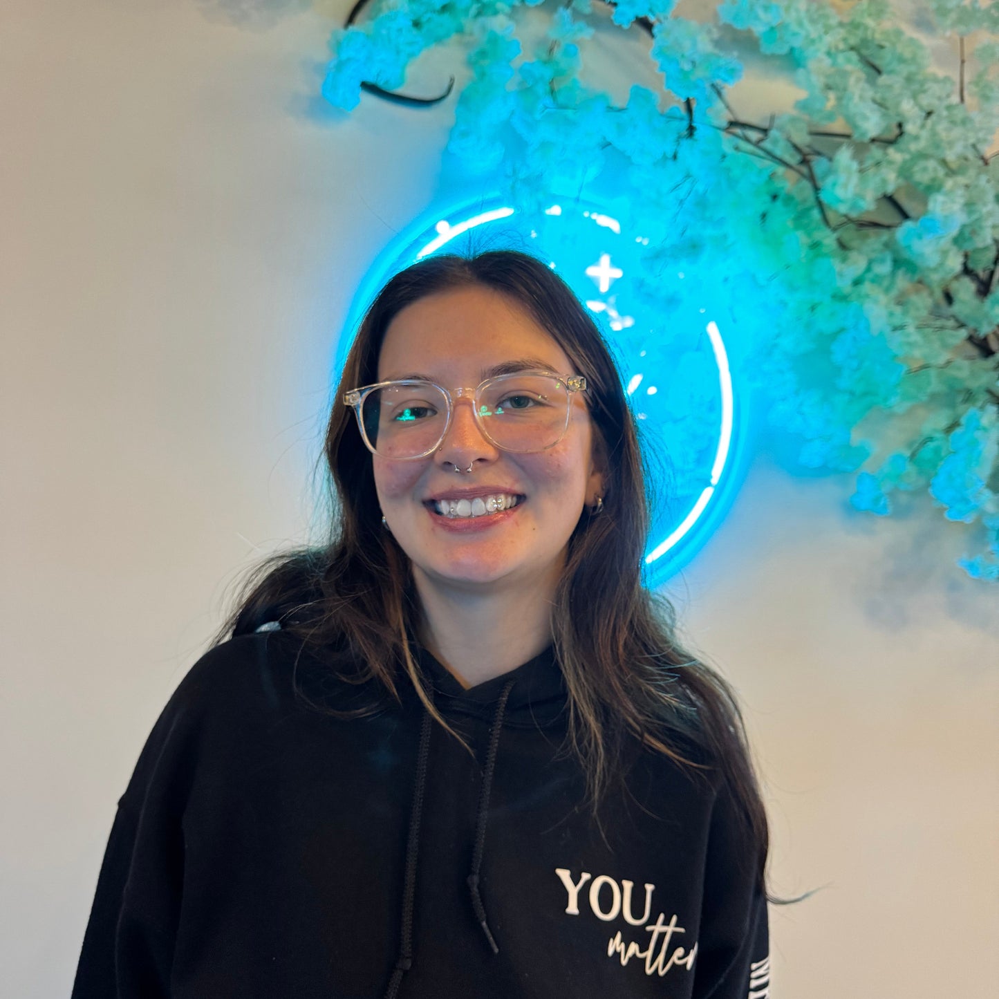 Person wearing glasses and a black hoodie with text at grin & glow, standing in front of a glowing blue circular light.