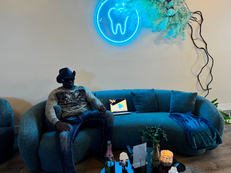 Person sitting on a blue couch with a neon tooth sign on the wall at Grin & Glow